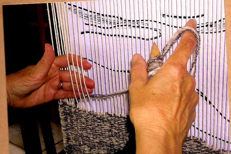 Gros plan des mains d'Aline Jegonday tissant à la flûte, le carton placé derrière les fils de chaîne